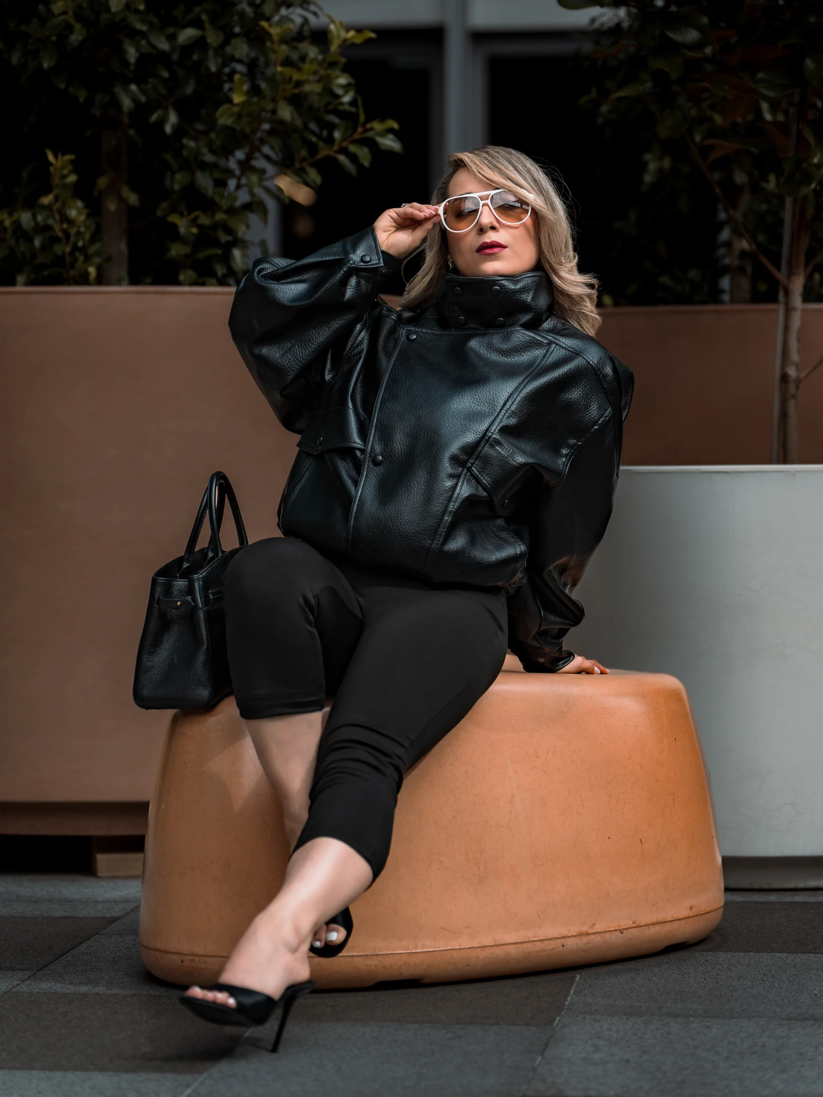 Neda seated in a black jacket and capri trousers, adjusting her white sunglasses