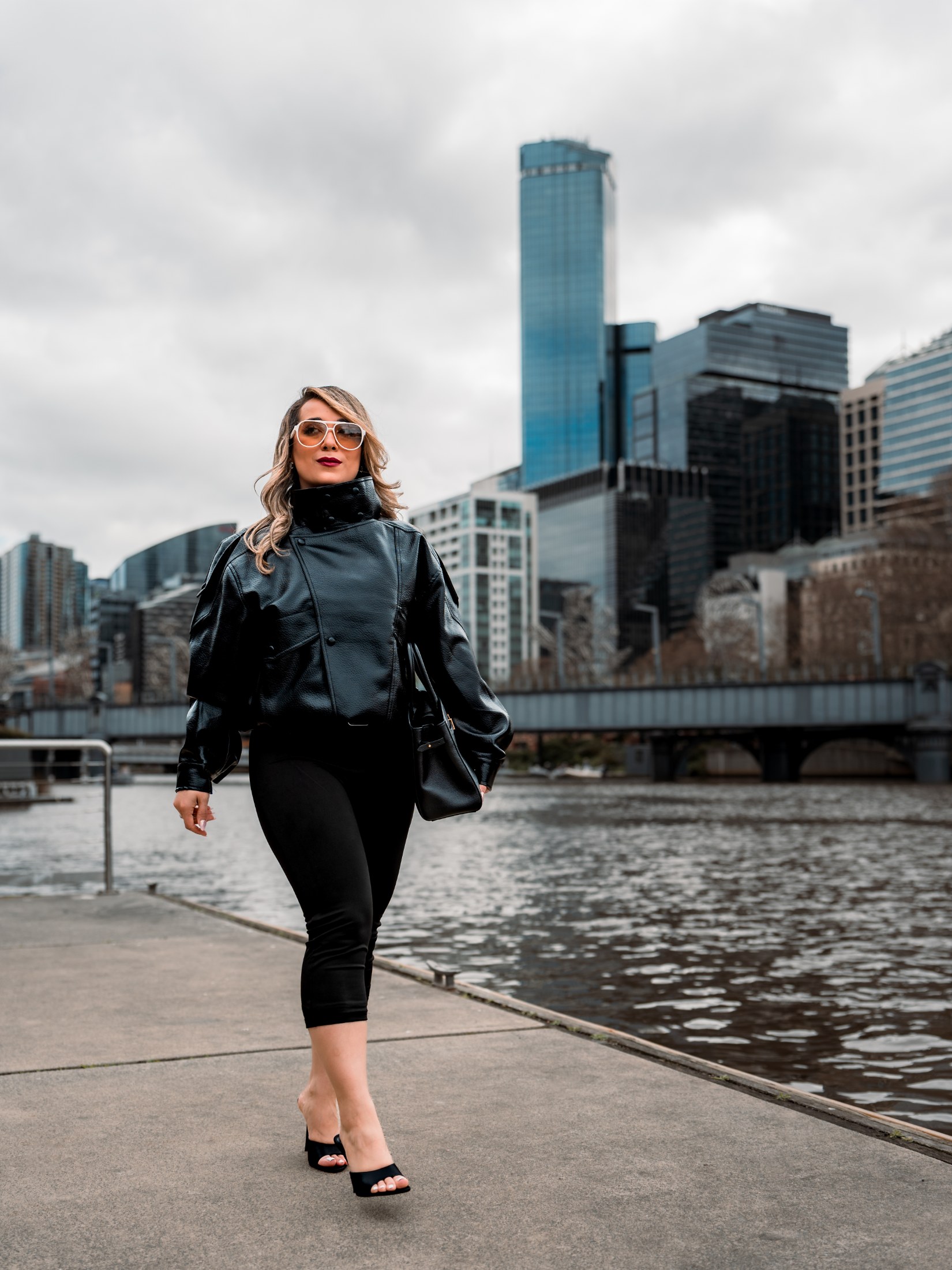 Neda wearing an all-black outfit beside the Yarra River with Melbourne’s skyline behind her